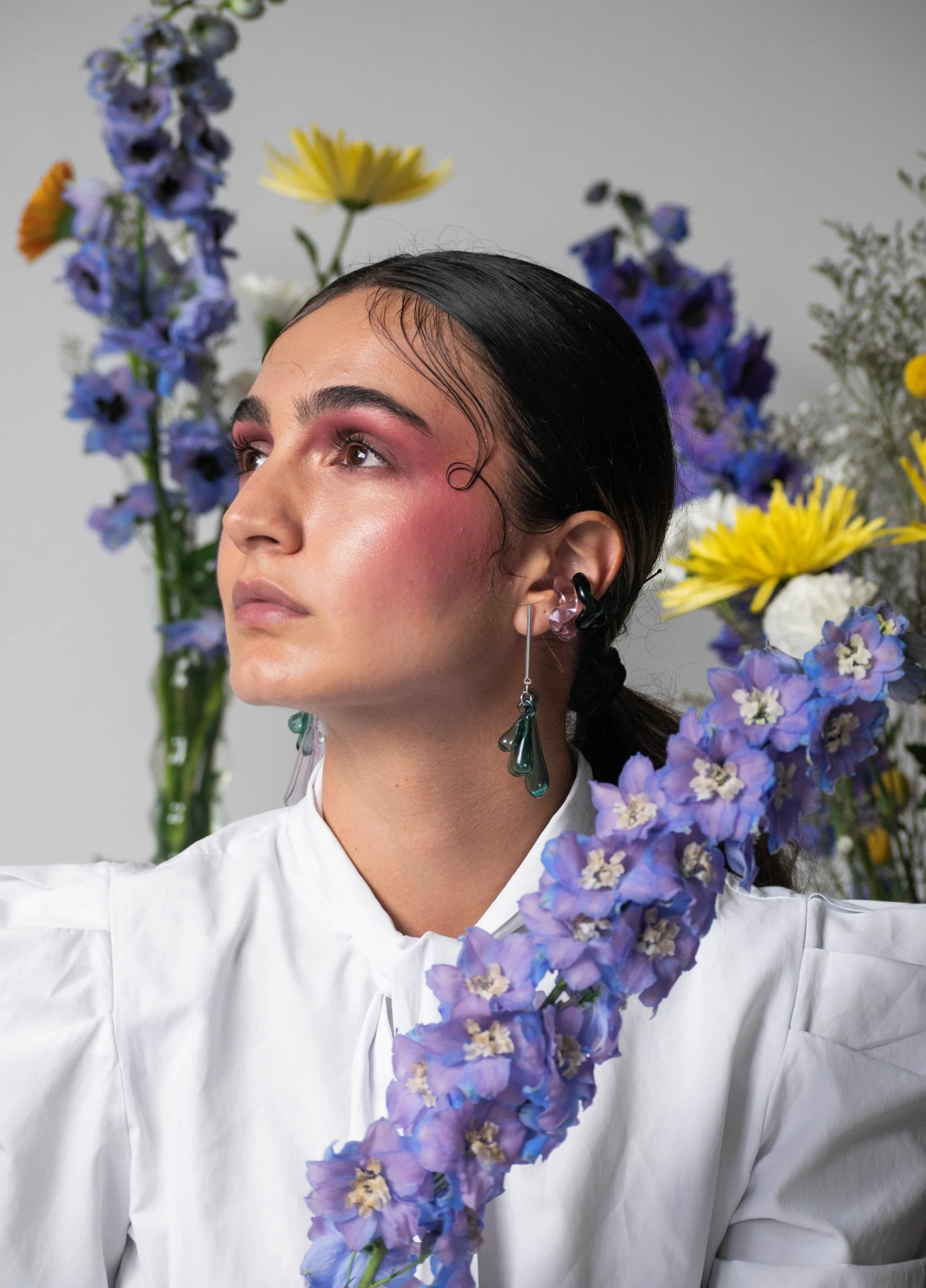 Three-quarter view portrait of person with artistic makeup and sculptural glass earrings, surrounded by purple delphiniums and yellow daisies