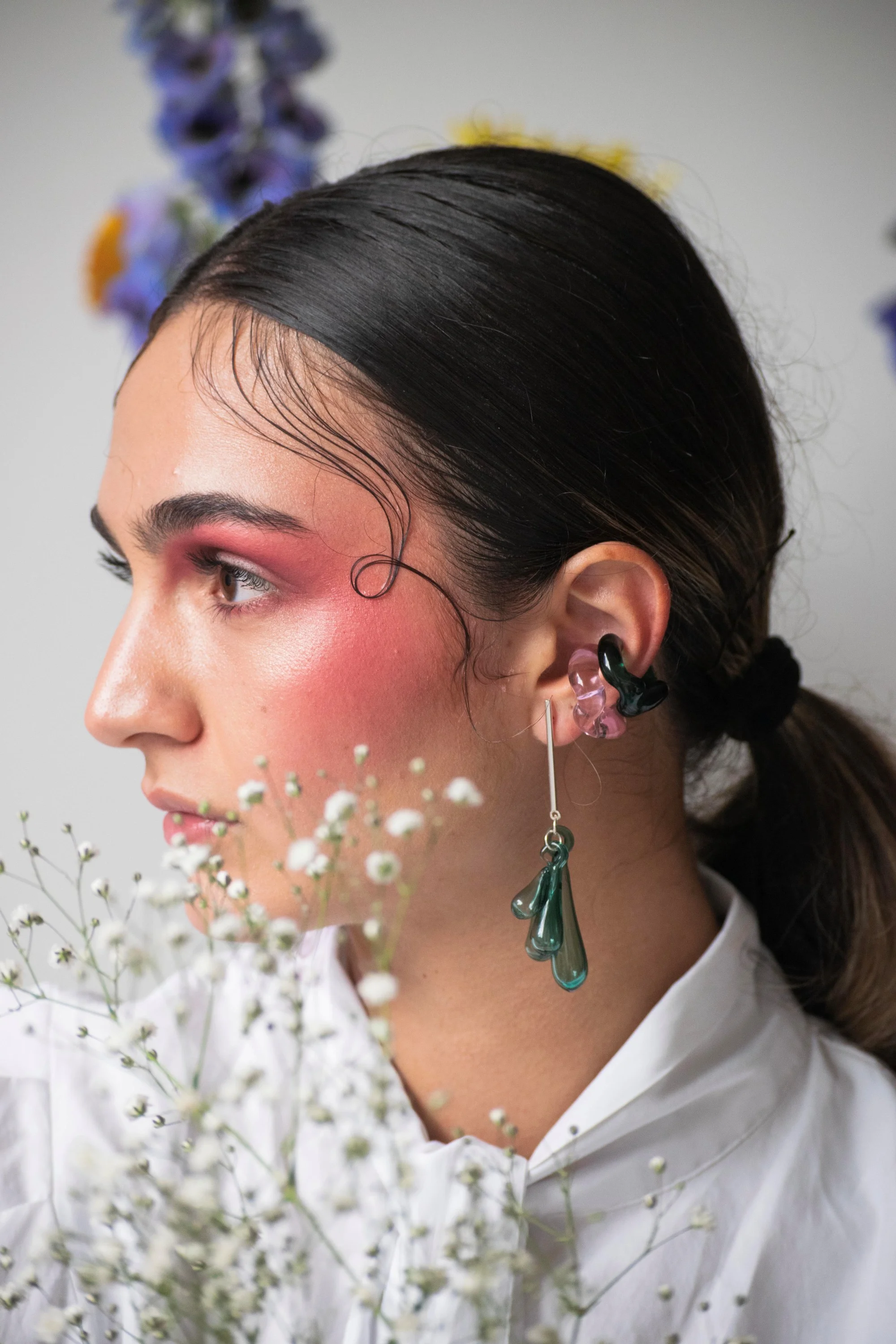 Profile portrait of person with slicked-back hair wearing artistic glass ear jewelry and pink gradient eye makeup, with delicate white flowers in foreground