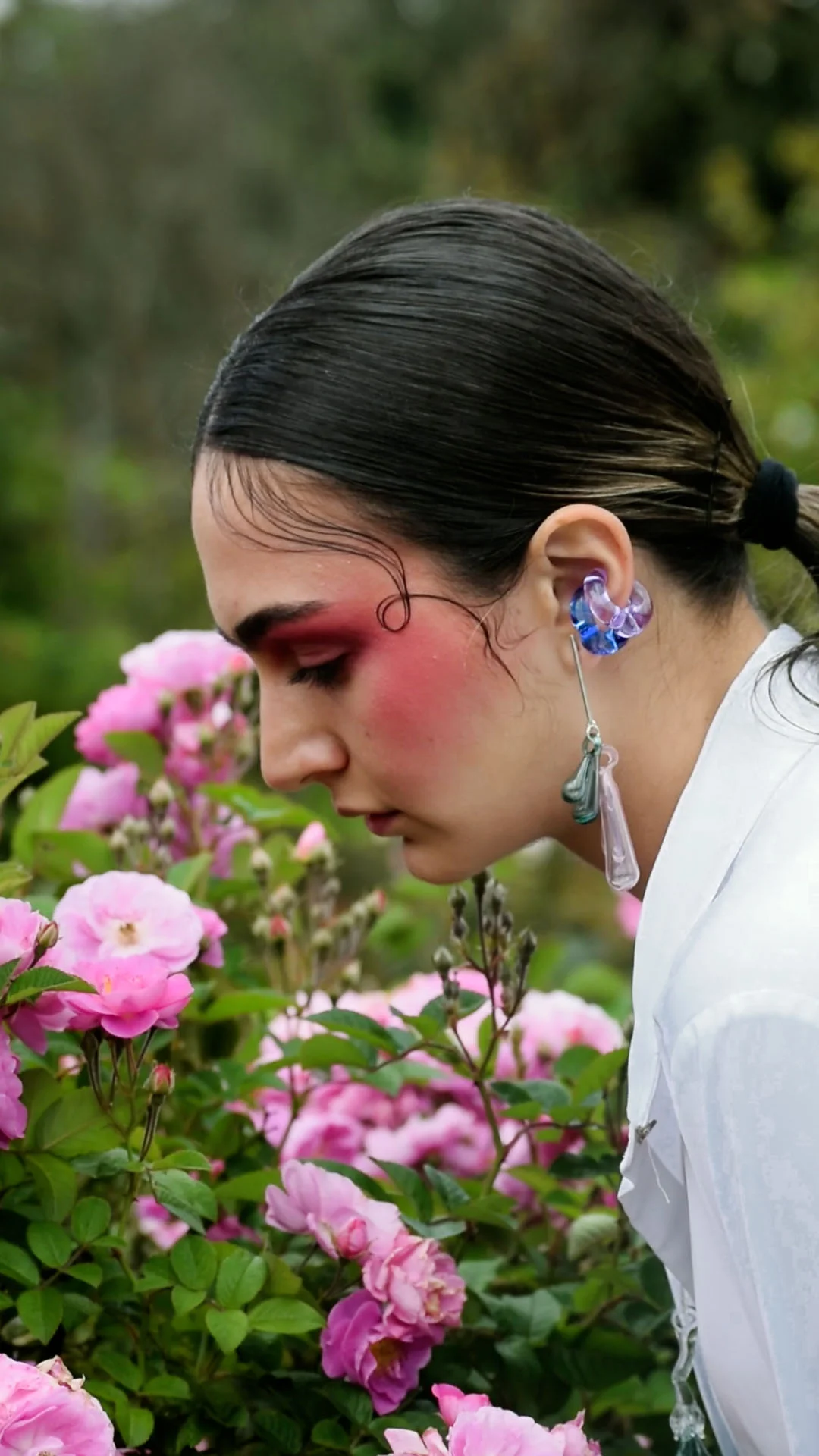 Side profile of person with artistic red eye makeup and transparent glass jewelry, leaning toward pink roses in natural garden setting