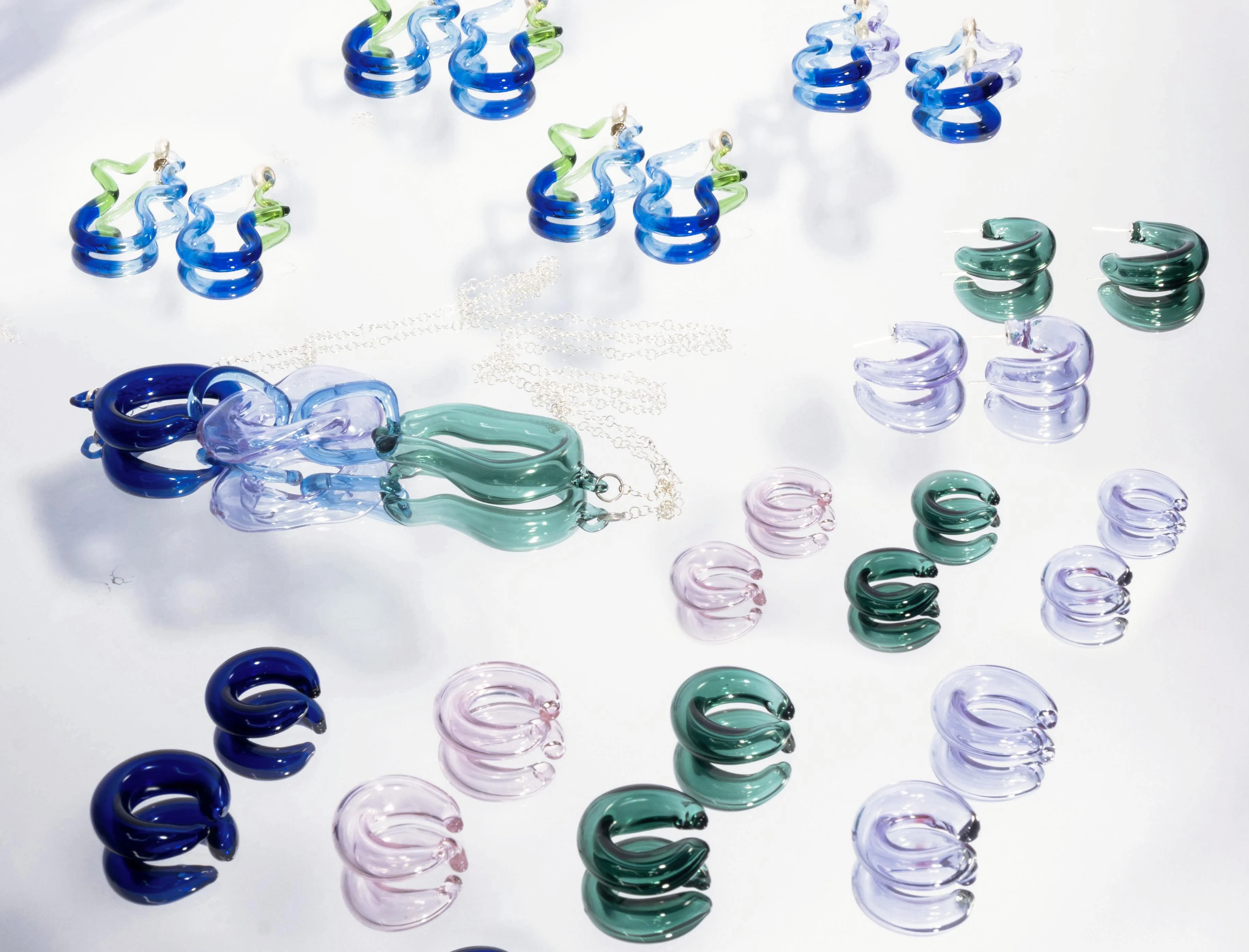 Vista cenital plana de colección de joyería escultórica de vidrio incluyendo ear cuffs, anillos y piezas con eslabones de cadena dispuestos sobre fondo blanco, con tonos azul cobalto, lavanda, verde esmeralda y rosa con acentos de cadena plateada