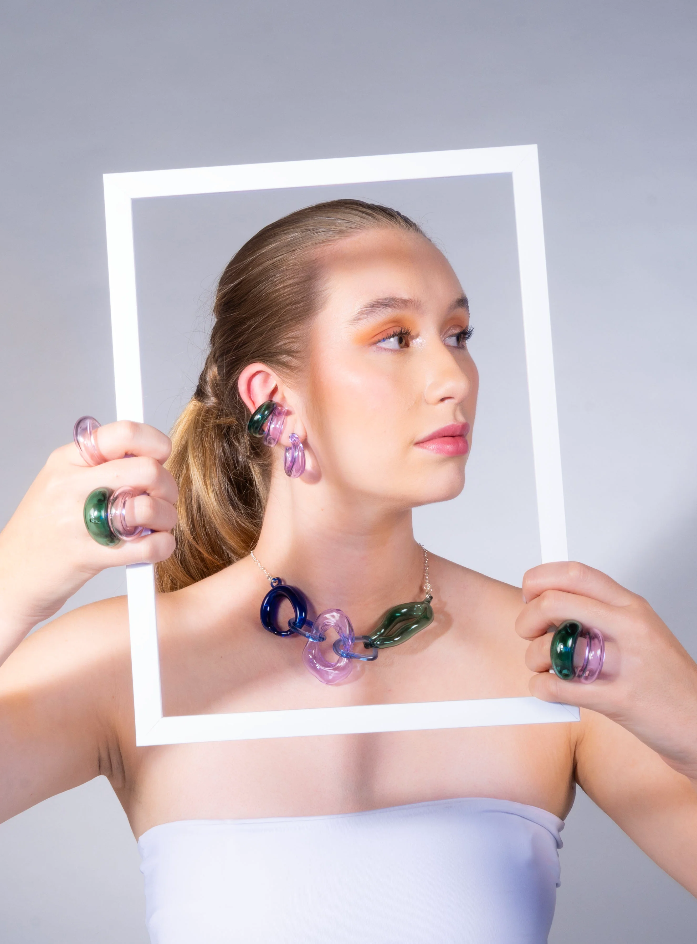 Modelo usando set completo de joyería incluyendo ear cuffs escultóricos de vidrio, anillos y collar de eslabones en lavanda, azul cobalto y verde esmeralda, sosteniendo marco blanco con maquillaje naranja en los ojos