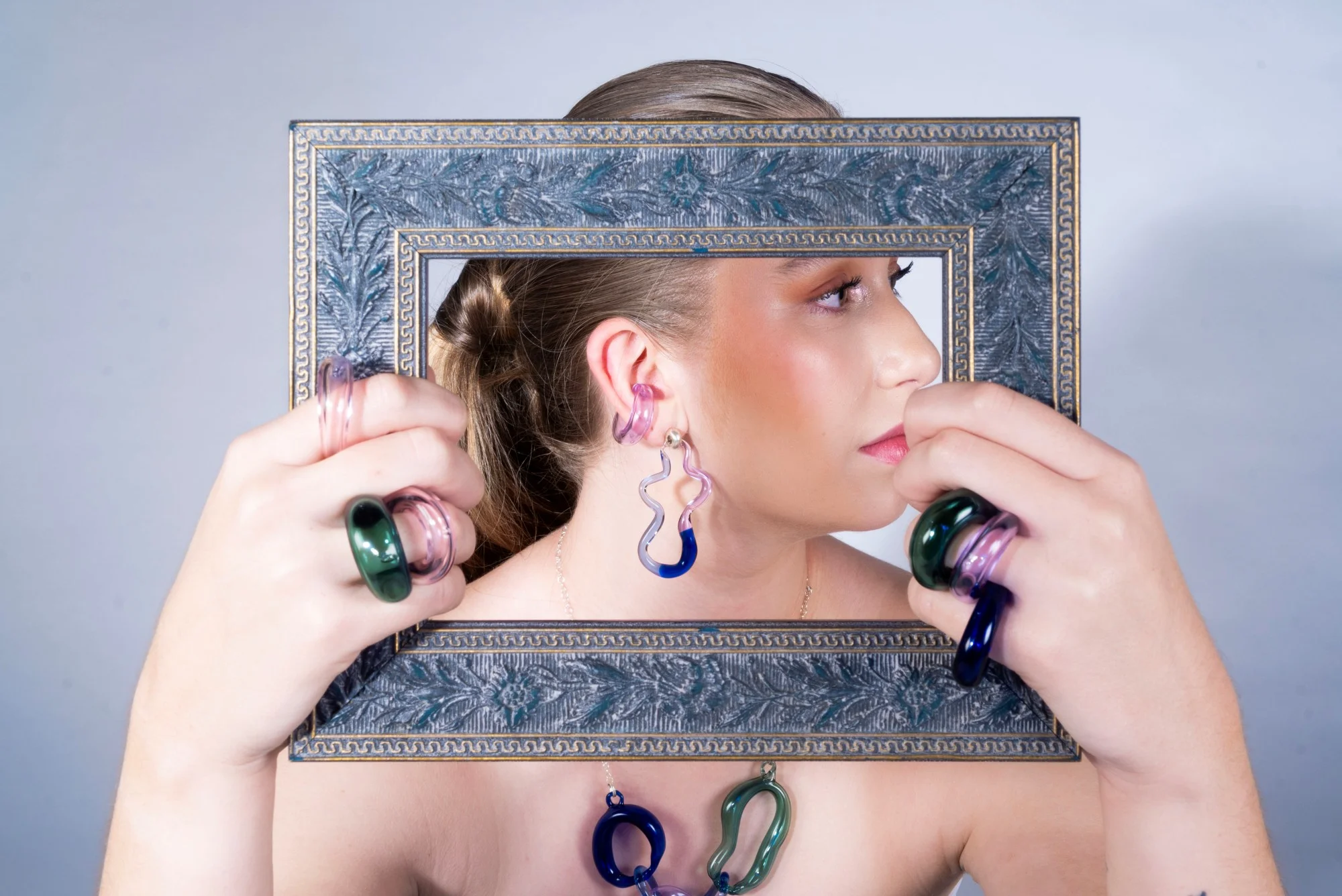 Retrato de vista trasera de modelo enmarcada por marco vintage ornamentado, usando joyería escultórica de vidrio incluyendo aretes ondulados colgantes, ear cuffs en espiral y collar de eslabones en rosa, azul cobalto y verde esmeralda