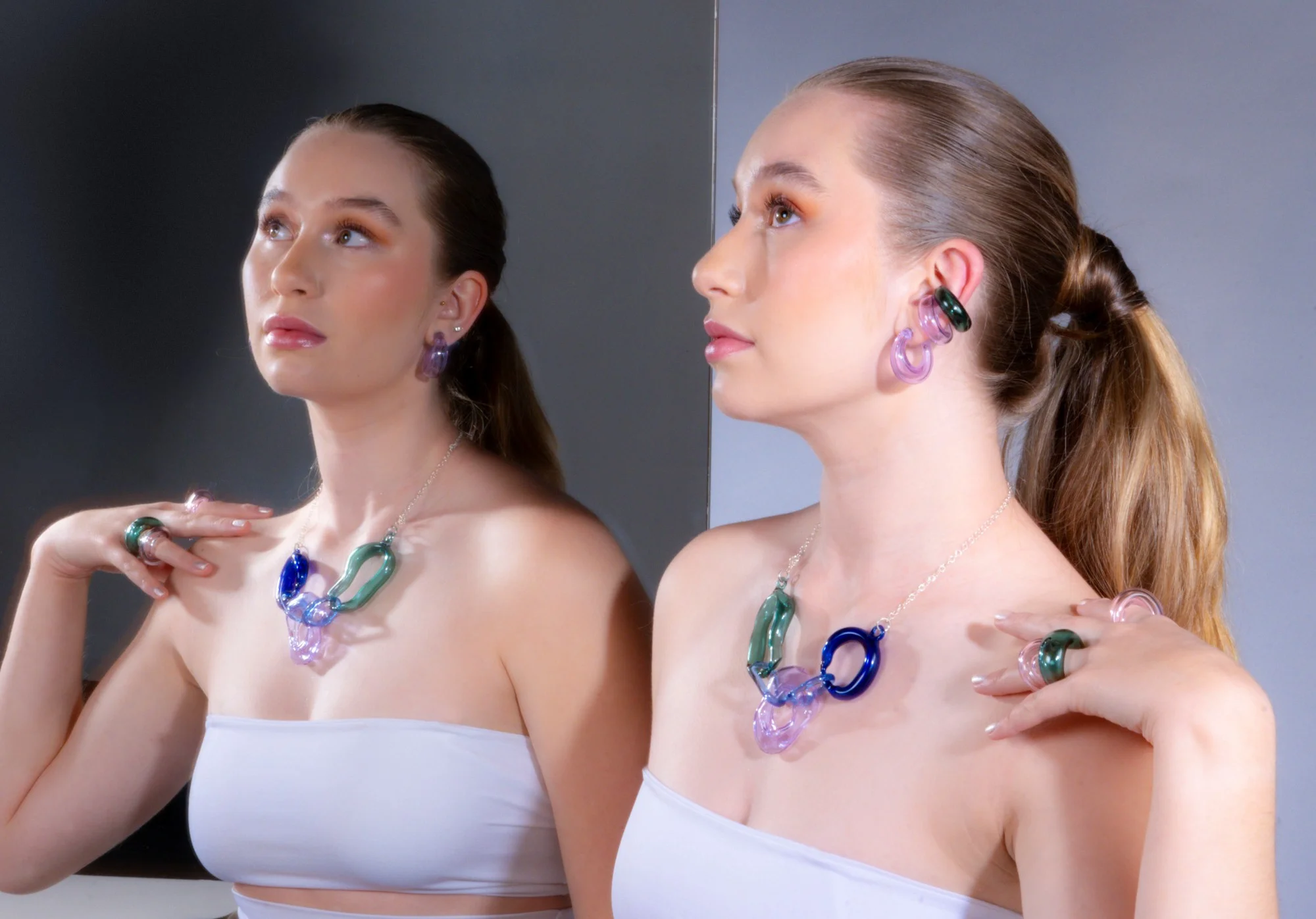Retrato dual mostrando modelo con cabello peinado hacia atrás usando set de joyería abstracta de vidrio, incluyendo ear cuffs, collar de eslabones y anillos en lavanda, azul cobalto y verde esmeralda contra fondo gris dividido
