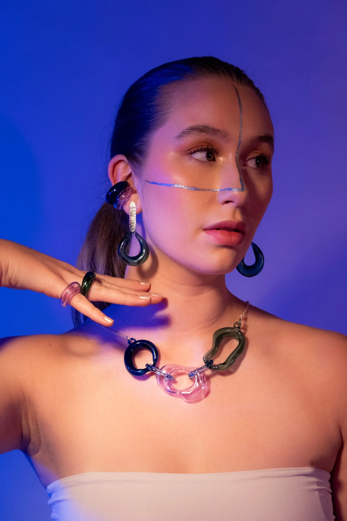 Retrato de moda con iluminación azul y naranja mostrando modelo usando joyería escultórica audaz de vidrio incluyendo aretes orgánicos colgantes, collar de eslabones con formas abstractas y anillos a juego en azul marino, rosa y verde