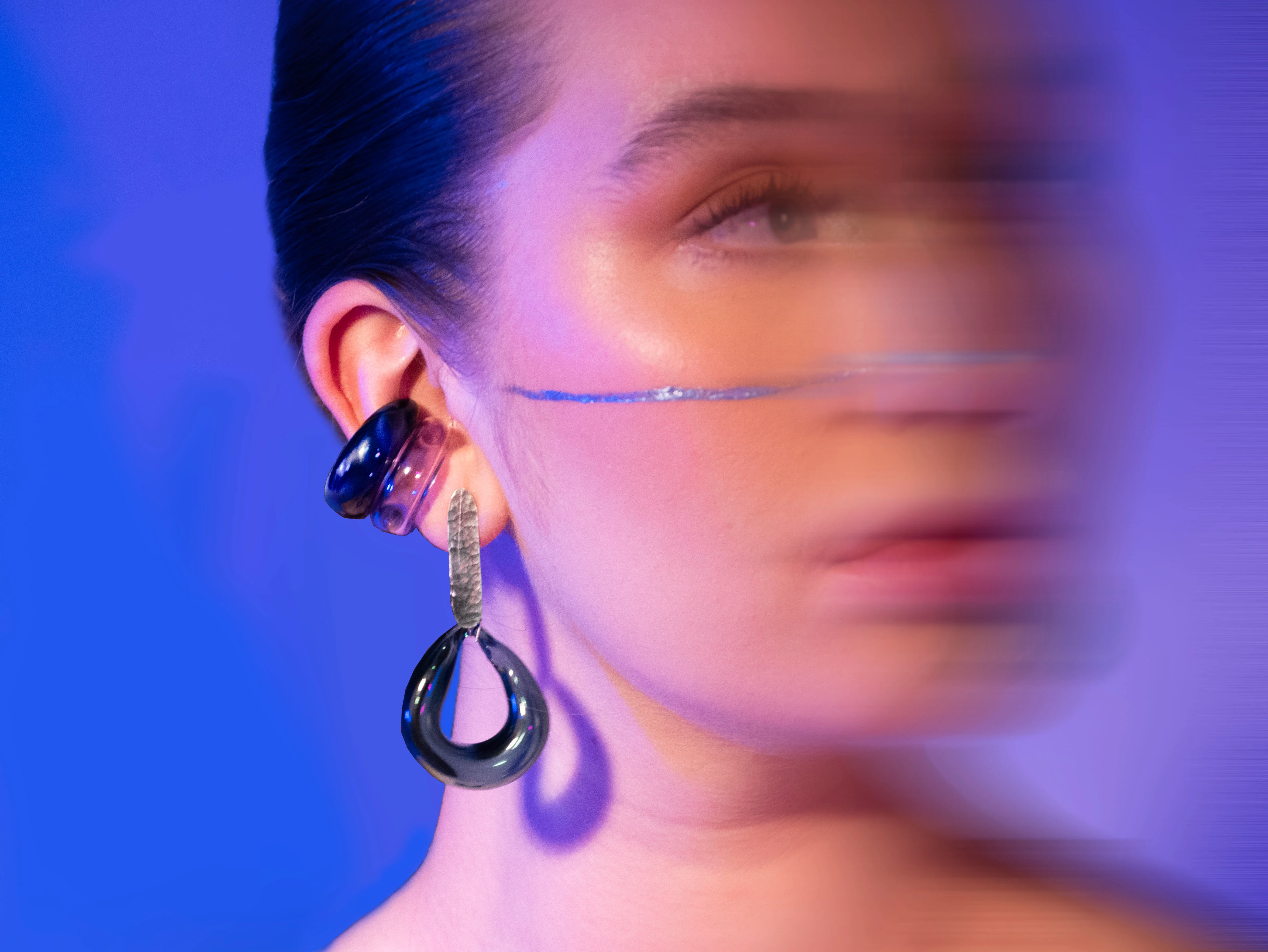 Close-up detail shot with motion blur showing sculptural glass ear cuff and drop earring with silver chain accent in navy blue and pink gradient under blue and purple lighting
