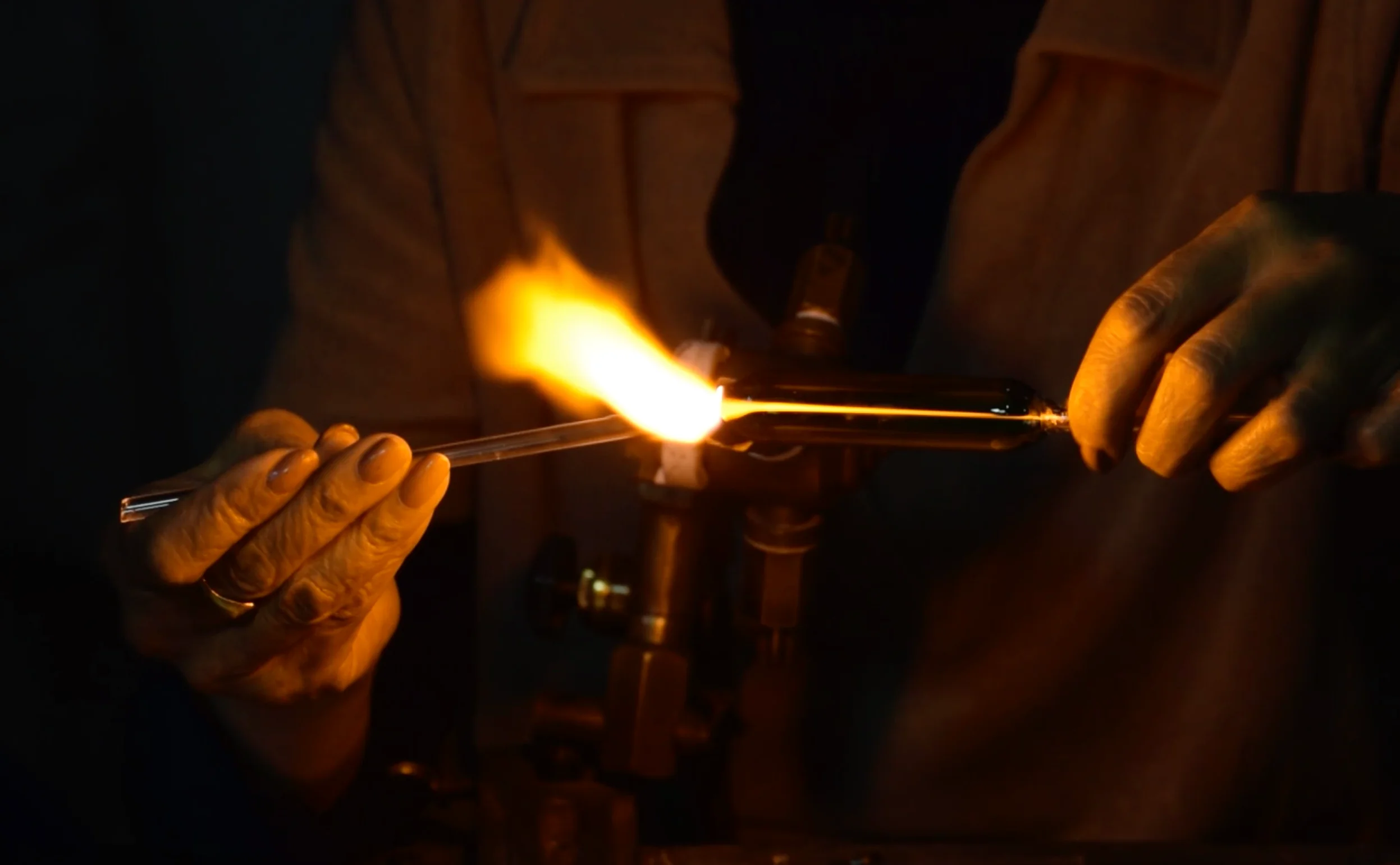 Manos de artesano trabajando vidrio caliente con soplete en un taller oscuro.