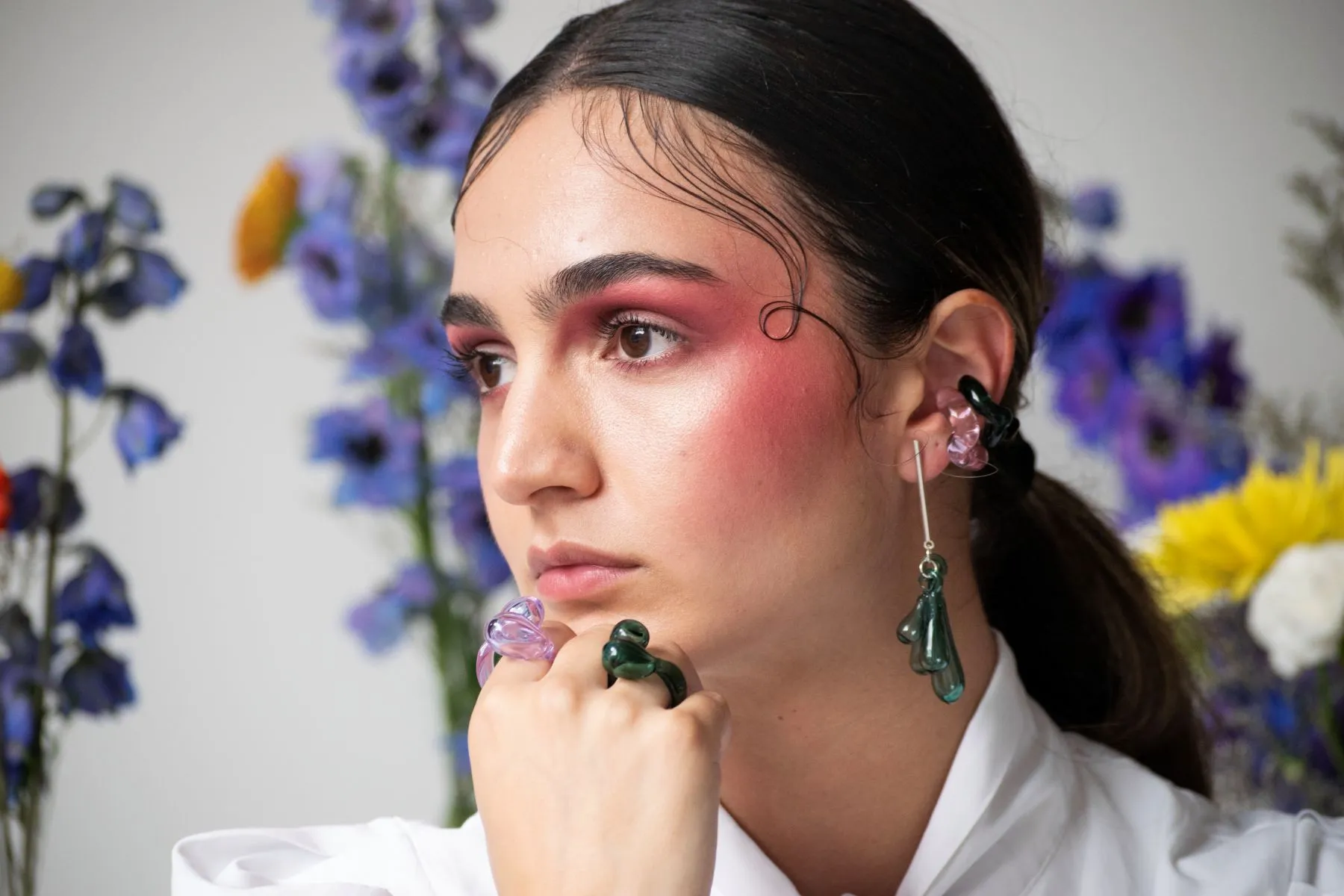 Retrato de una mujer con maquillaje rojo intenso usando joyería artística de resina, incluyendo anillos, aretes y ear cuff, con fondo floral.