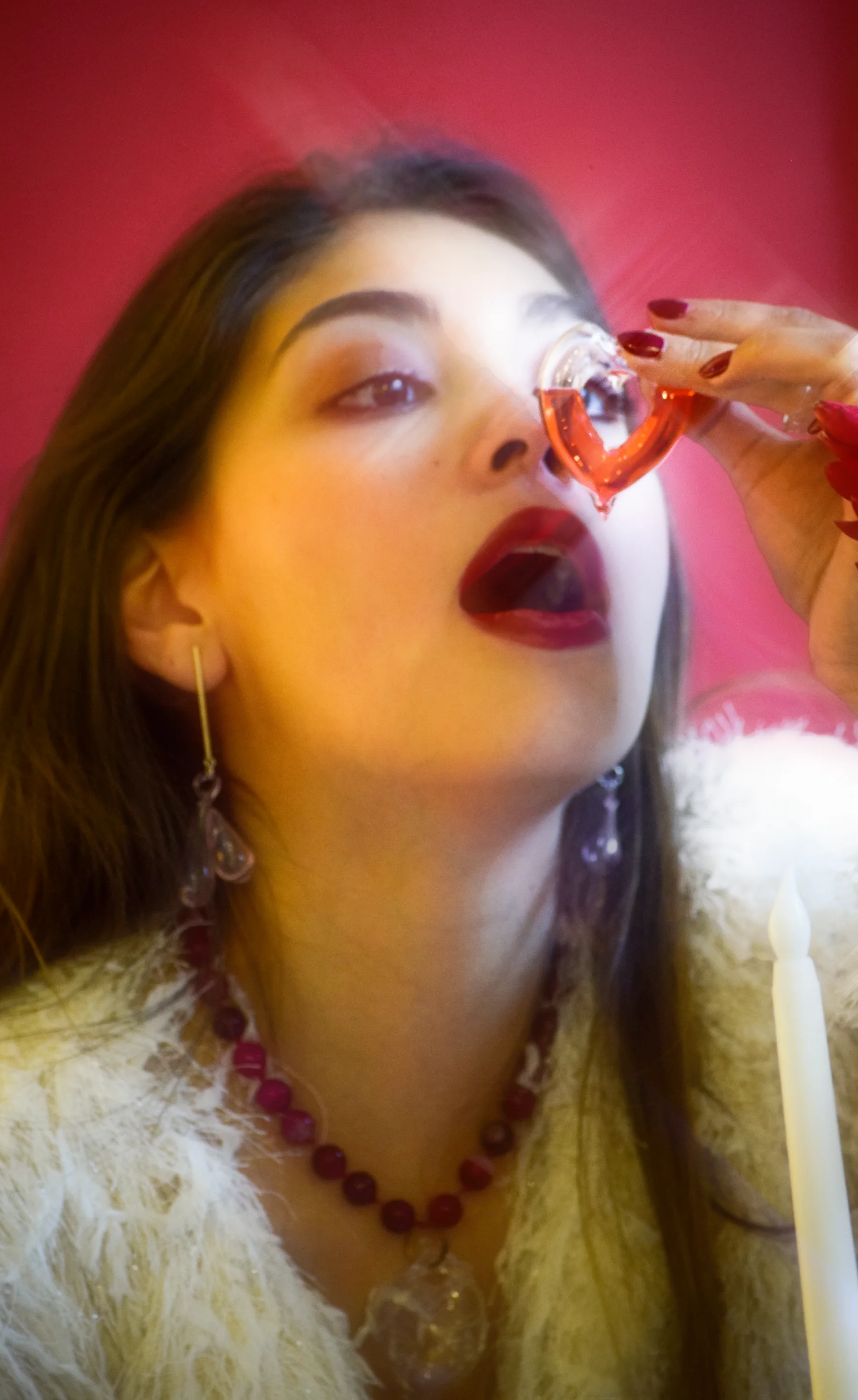 Primer plano de una mujer con maquillaje rojo intenso sosteniendo una pieza translúcida en forma de corazón cerca de su ojo, usando joyería artística y un collar de cuentas, con fondo rojo vibrante.