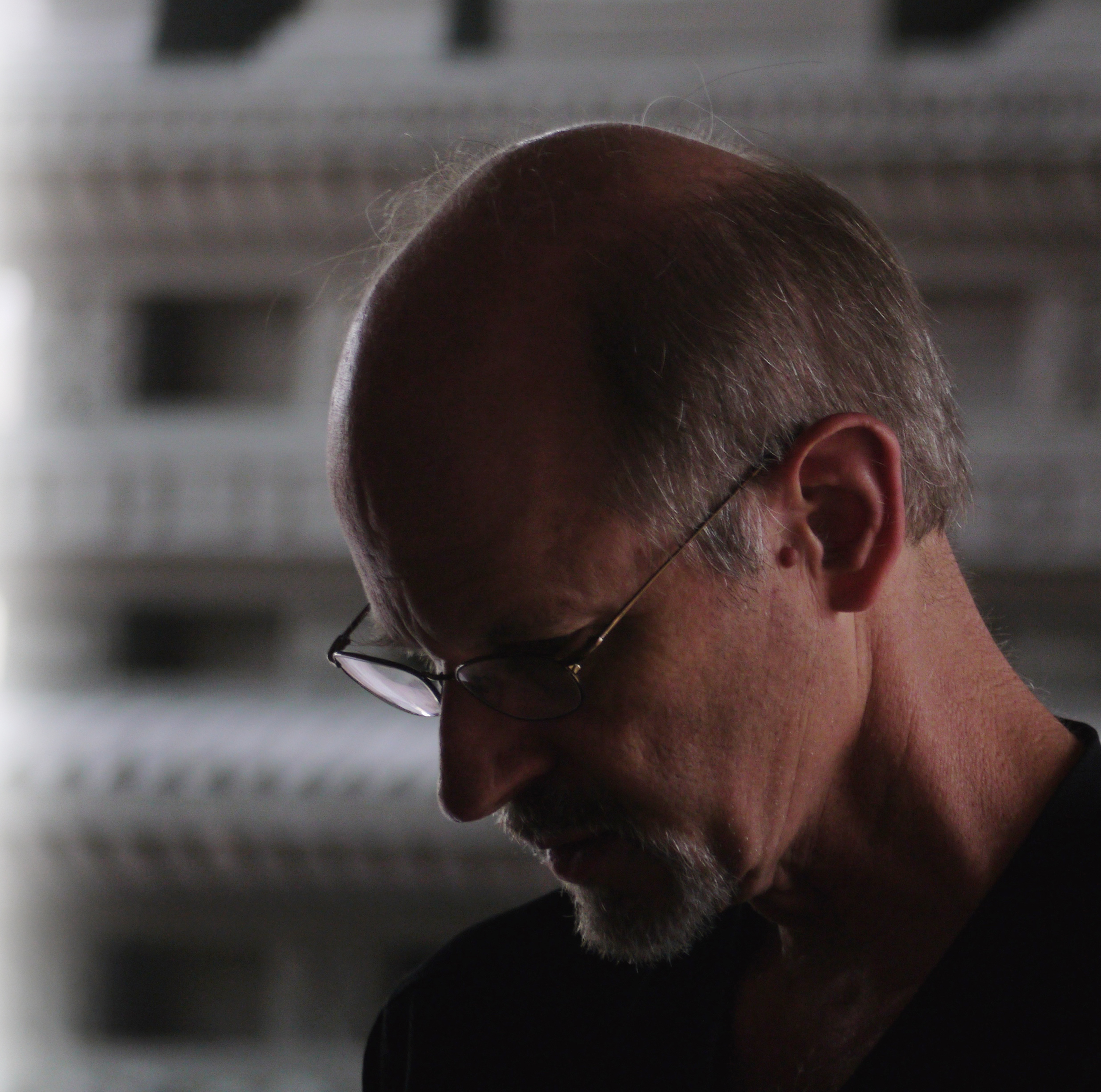 Close-up profile of an older man with glasses looking down thoughtfully in a dark setting.