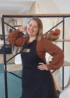 Біля декоративної стінки стоїть і посміхається Ірина - власниця кавʼярні. Одягнута у коричневу сорочку в клітинку та чорний фартук з логотипом Наша Кава