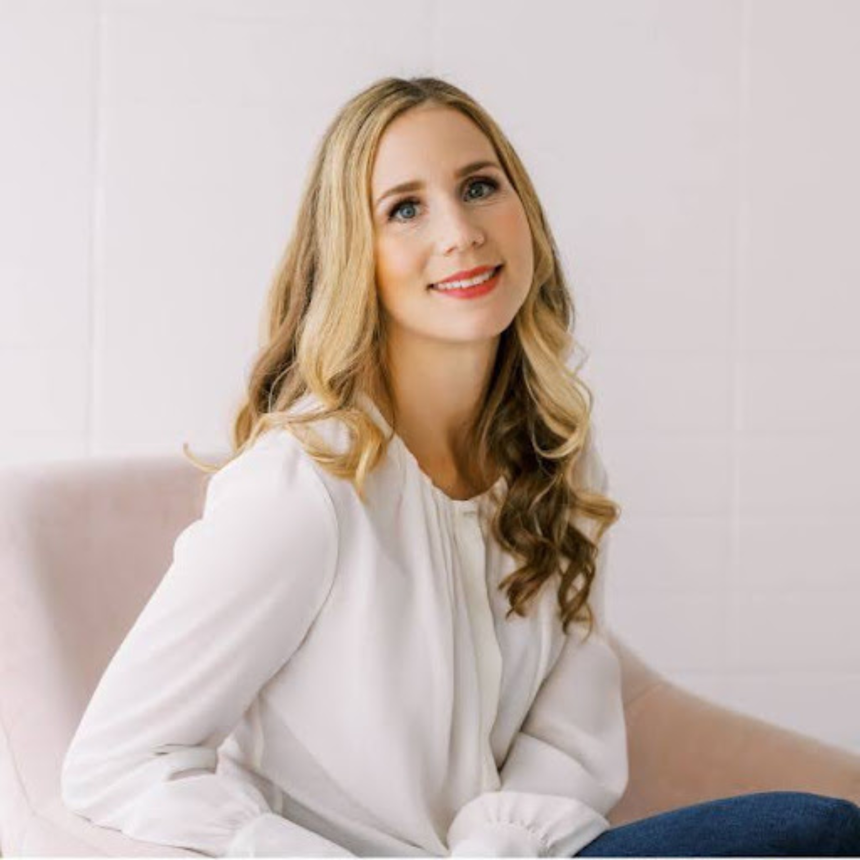 Smiling woman with long wavy blonde hair looking upward from a soft pink chair against a warm peach background.