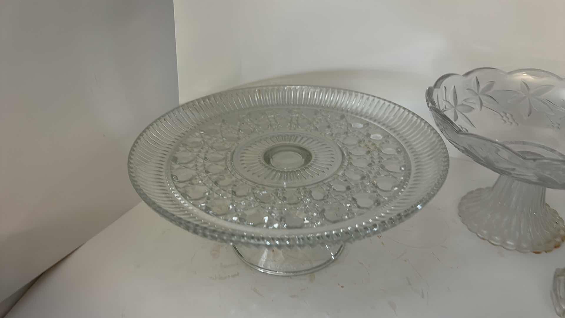Photo 1 of ASSORTED VINTAGE CLEAR PRESSED GLASS SERVING PIECES INCLUDING DECORATIVE ROUND PLATTER, PEDESTAL COMPOTE AND FLORAL RELIEF DISH.
ESTIMATED RETAIL VALUE: $40–$60 FOR THE GROUP.