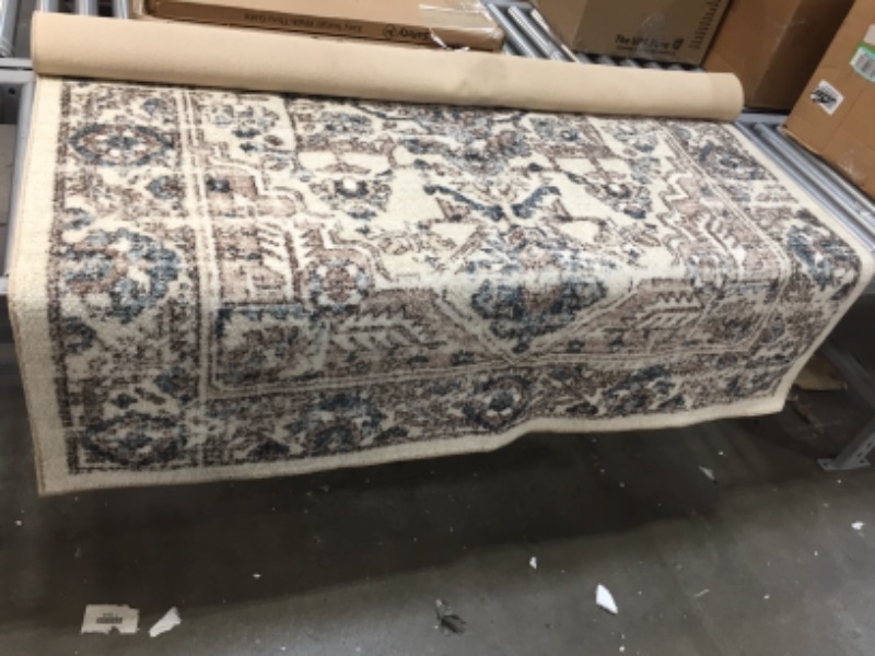 Photo 1 of **UNKNOWN BRAND**GENERAL POST**RUBBER ON BOTTOM IS VISIBLY WORN OUT**
5'X7' BEIGE RUG WITH BLUE AND BROWN
