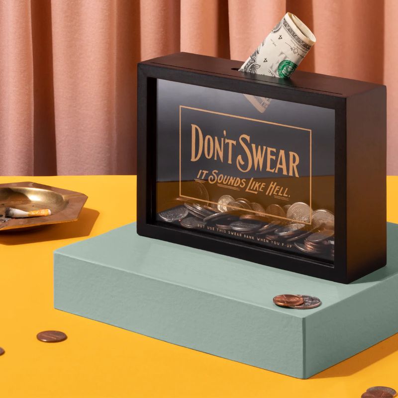Photo 1 of Don't Swear Swear Bank. Whoever said that swearing doesn't pay off? So do what you love, and save money all at the same time. This swear bank by Brass Monkey features a glass front (with gold foil art) and a slot for coins on the top. It's even f-ing hang