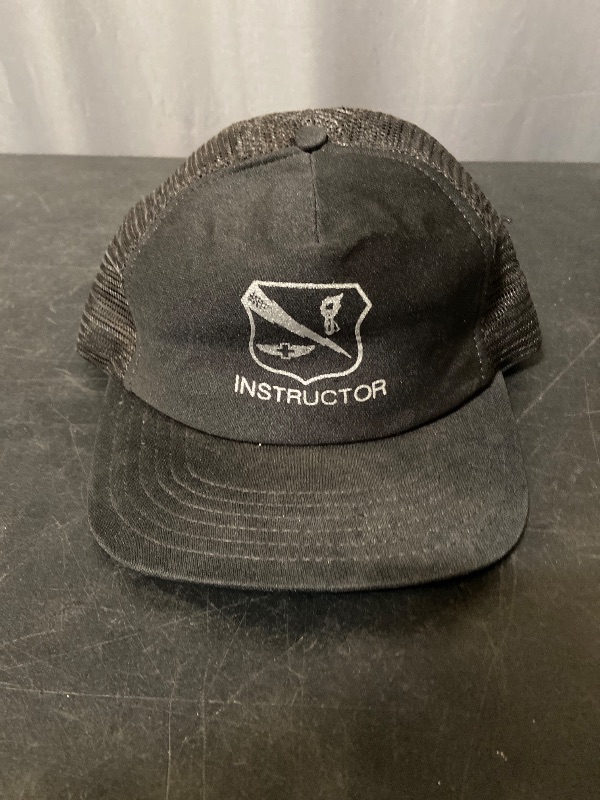 Photo 1 of vintage black trucker hat features a white embroidered "INSTRUCTOR" patch with an aviation-inspired logo.
