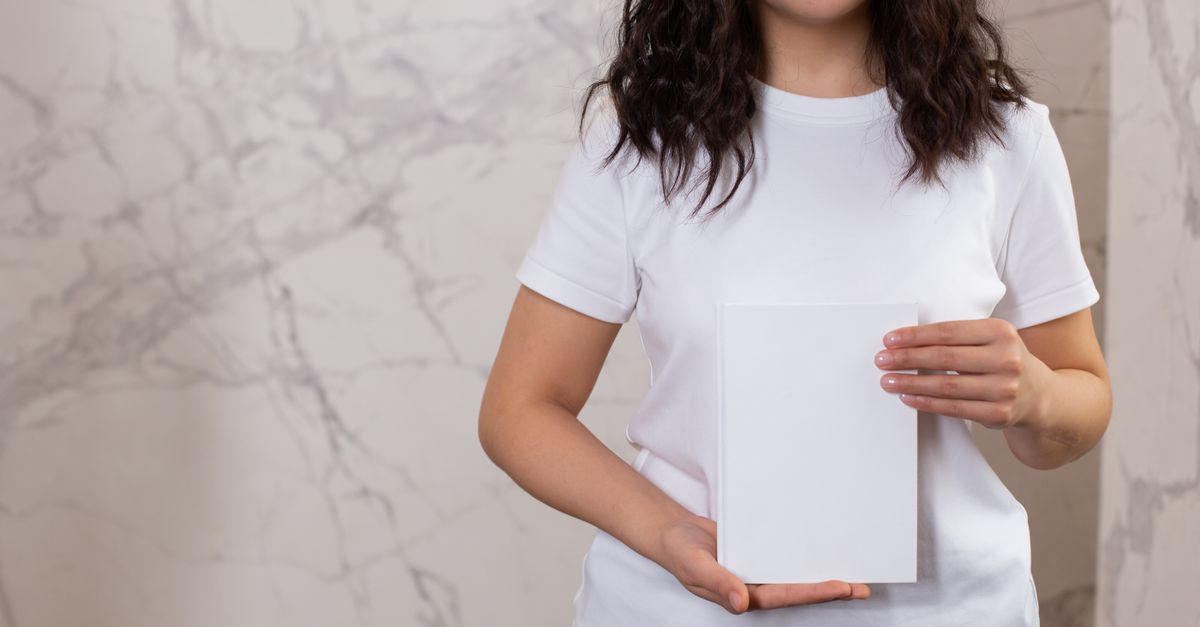 Unrecognizable female with black hair and casual t shirt showing white black notepad while standing near wall in light room