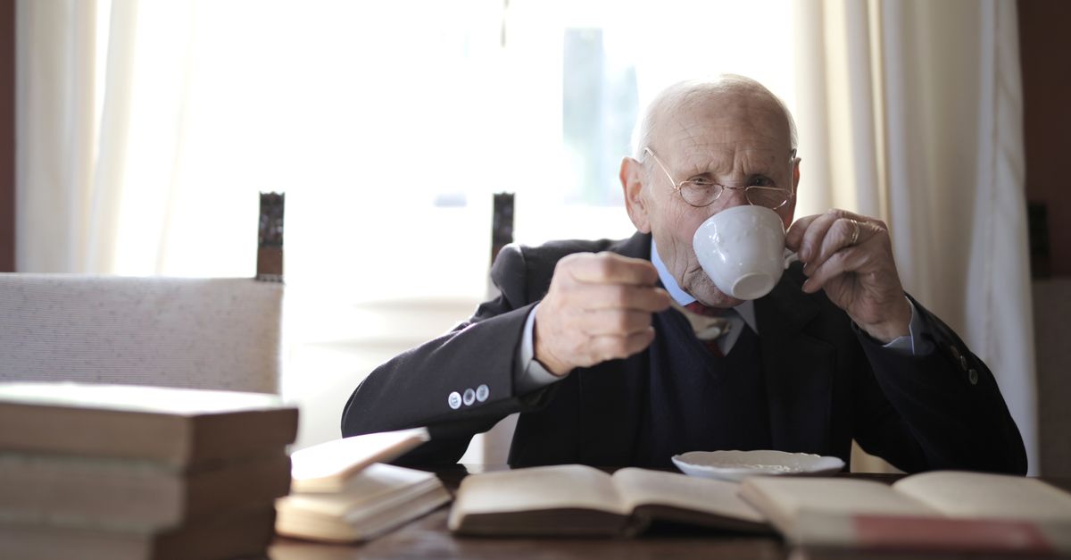 Senior man in formal suit and eyeglasses drinking tea from cup while holding spoon in hand and sitting at table with books