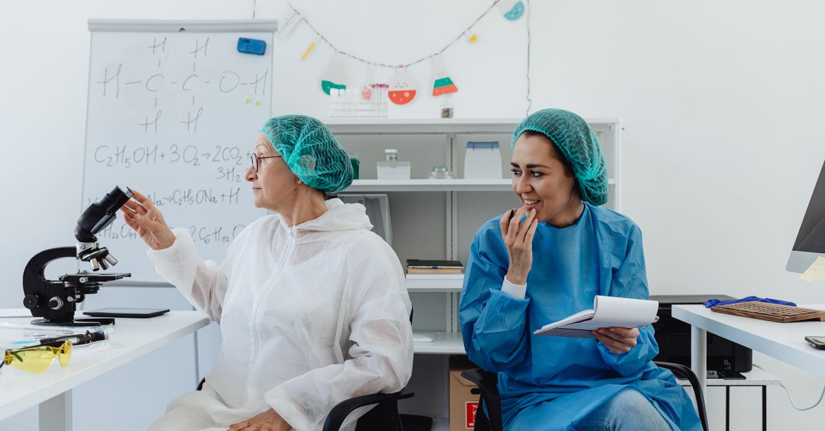 Two women scientists, in protective gear, working in a modern laboratory setting with a microscope and notepad.