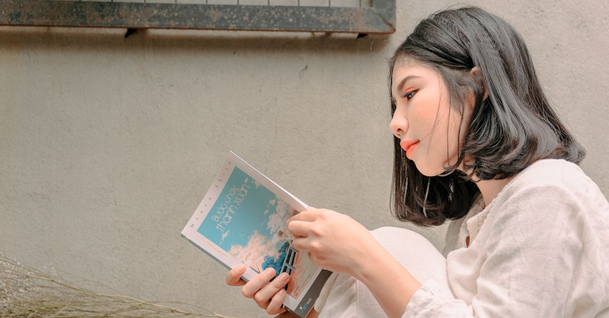 A young woman deeply engaged in reading a book while sitting outdoors, captured in natural daylight.
