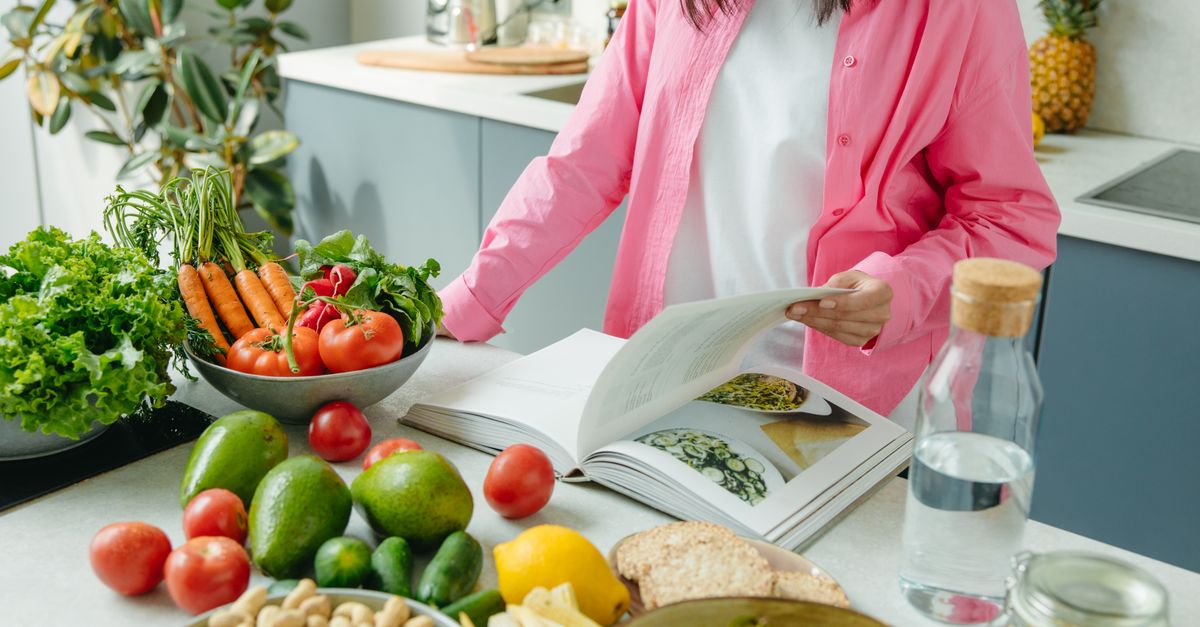 A person browsing a cookbook surrounded by fresh vegetables in a bright kitchen setting.