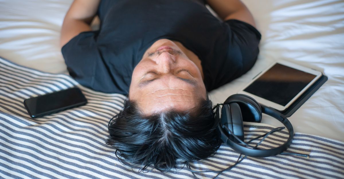 An adult man relaxing indoors with eyes closed, surrounded by gadgets like a smartphone, tablet, and headphones.