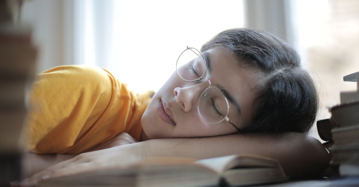 Young ethnic woman in eyeglasses and casual wear tired of exam preparation lying asleep on table with stacks of books