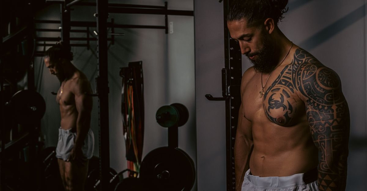 Shirtless muscular man with tattoos standing in a gym setting by the mirror.