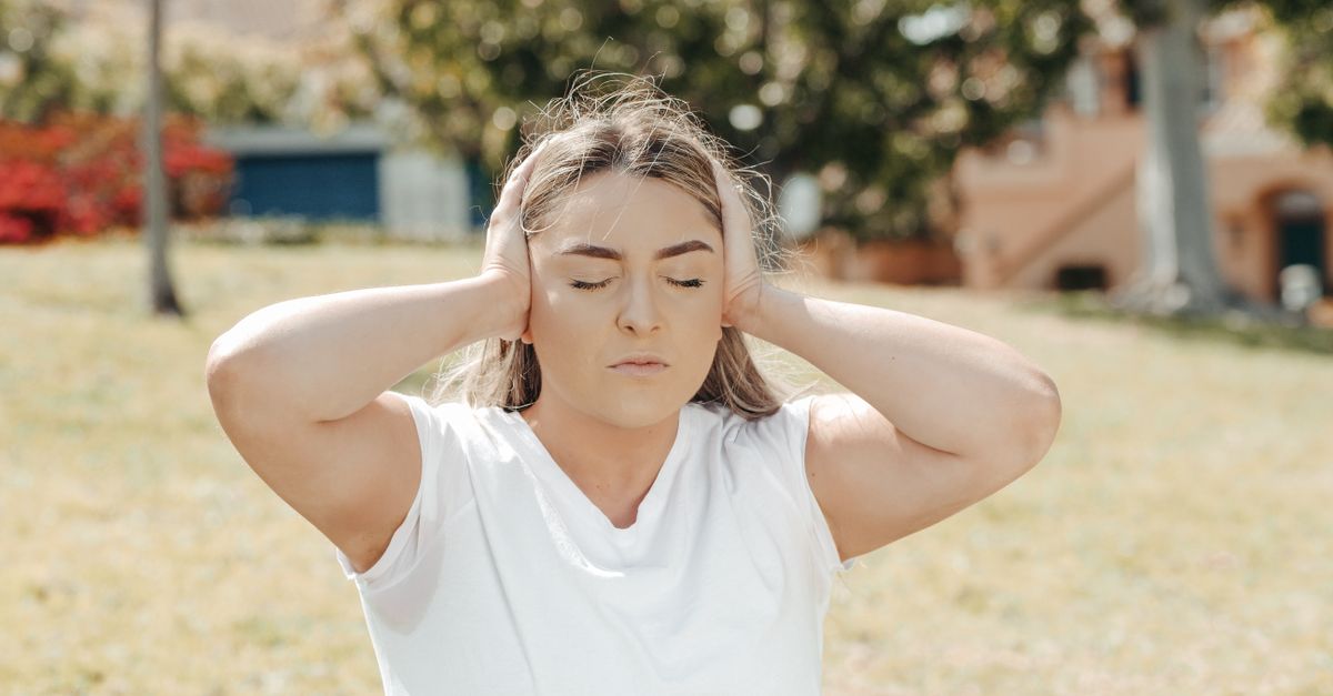 Caucasian woman in a park, eyes closed, holding head in pain hinting at a headache or migraine.
