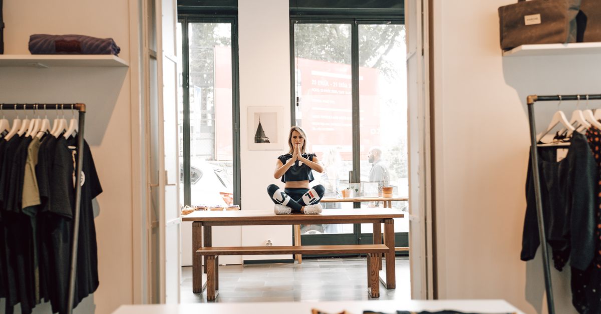 Stylish female in sportswear sitting with crossed legs during meditation and looking at camera on table in boutique with apparel on racks