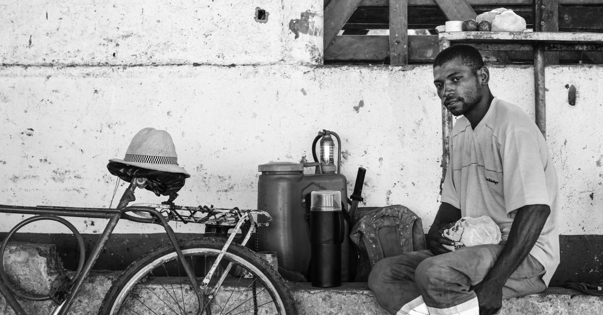 Black and white photo of a man sitting by a bicycle against a wall in Vassouras, Brazil.