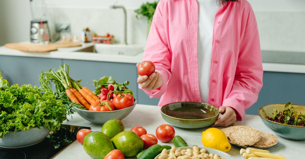 Woman in pink overshirt holds tomato in kitchen with fresh fruits and veggies.