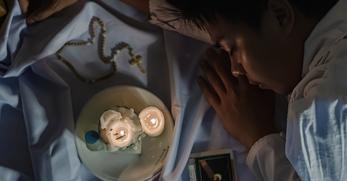 From above side view of crop religious Indian male sleeping on crumpled fabric with rosary and burning candles
