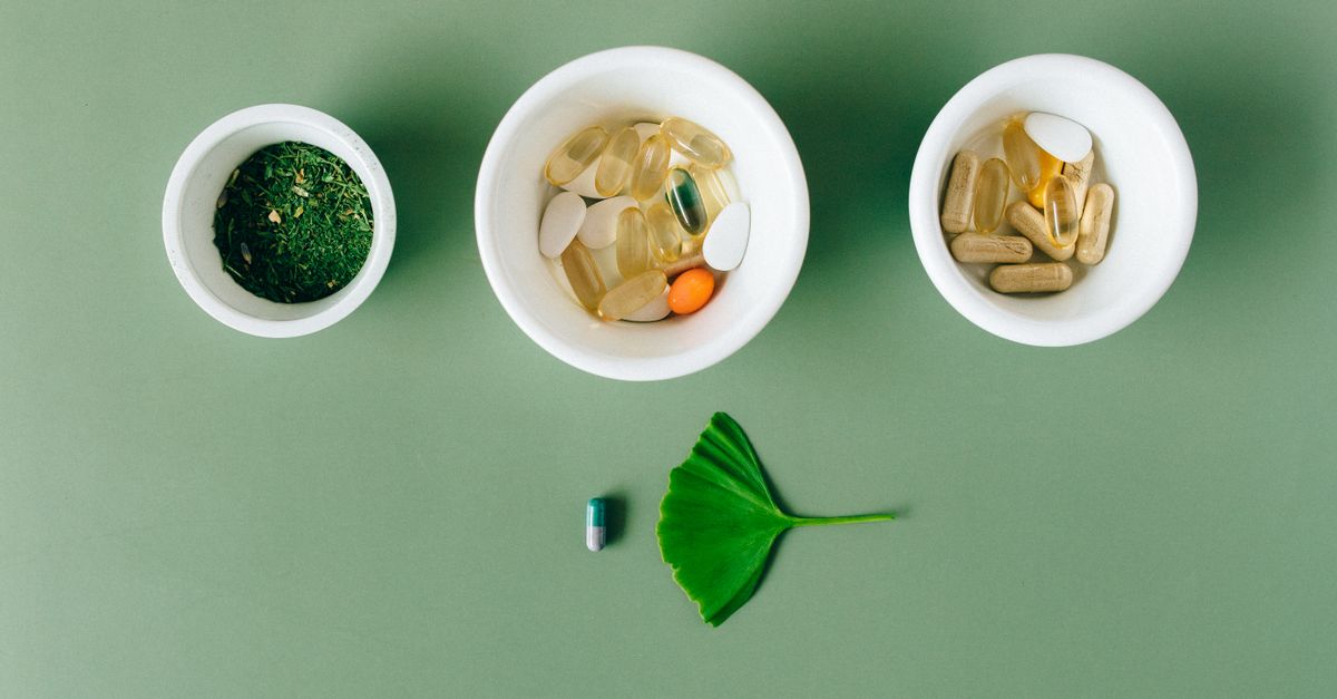 Flatlay of capsules and herbs in ceramic bowls on green surface.