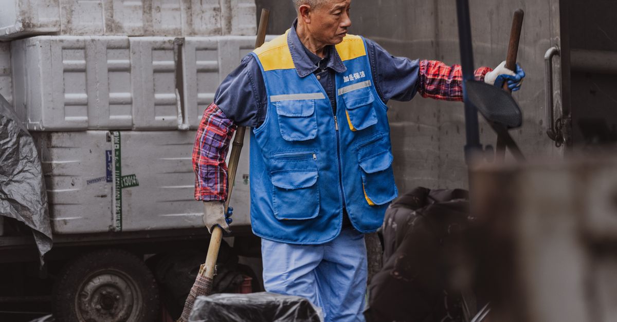 An adult male worker wearing a blue vest is engaged in outdoor work involving equipment handling.