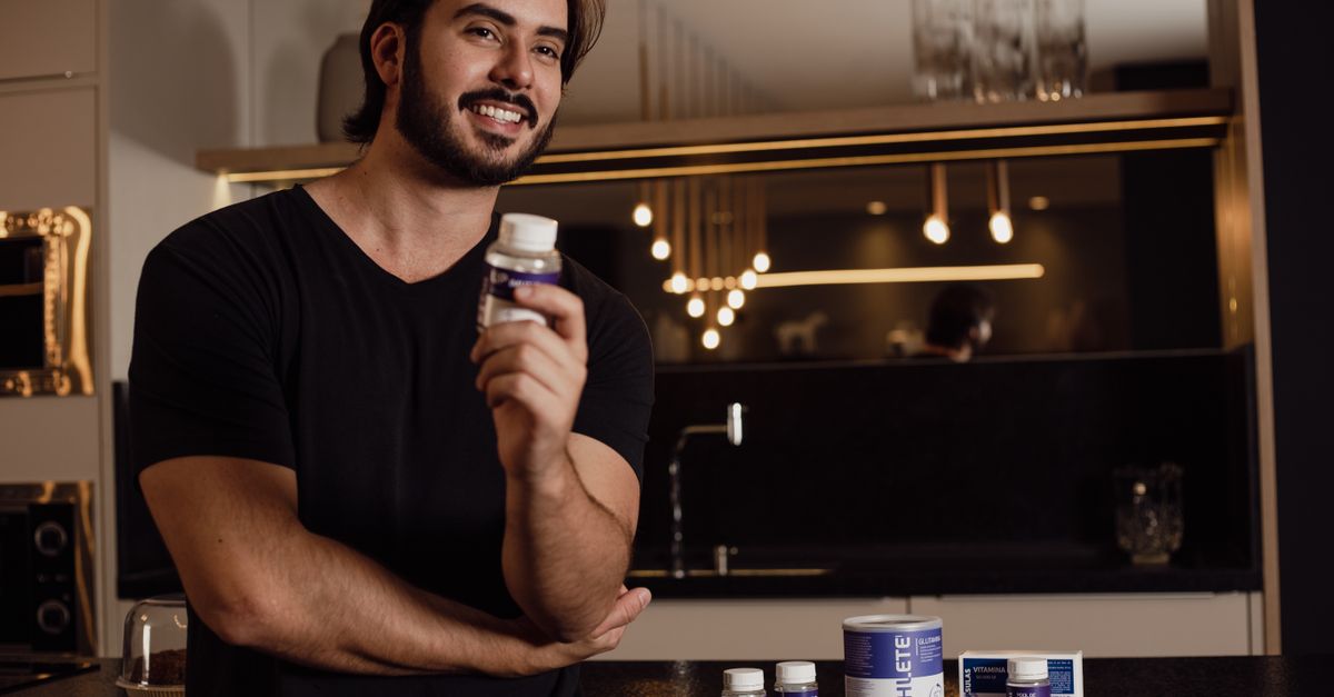 Smiling man holding a supplement bottle in an indoor kitchen setting with warm lighting.