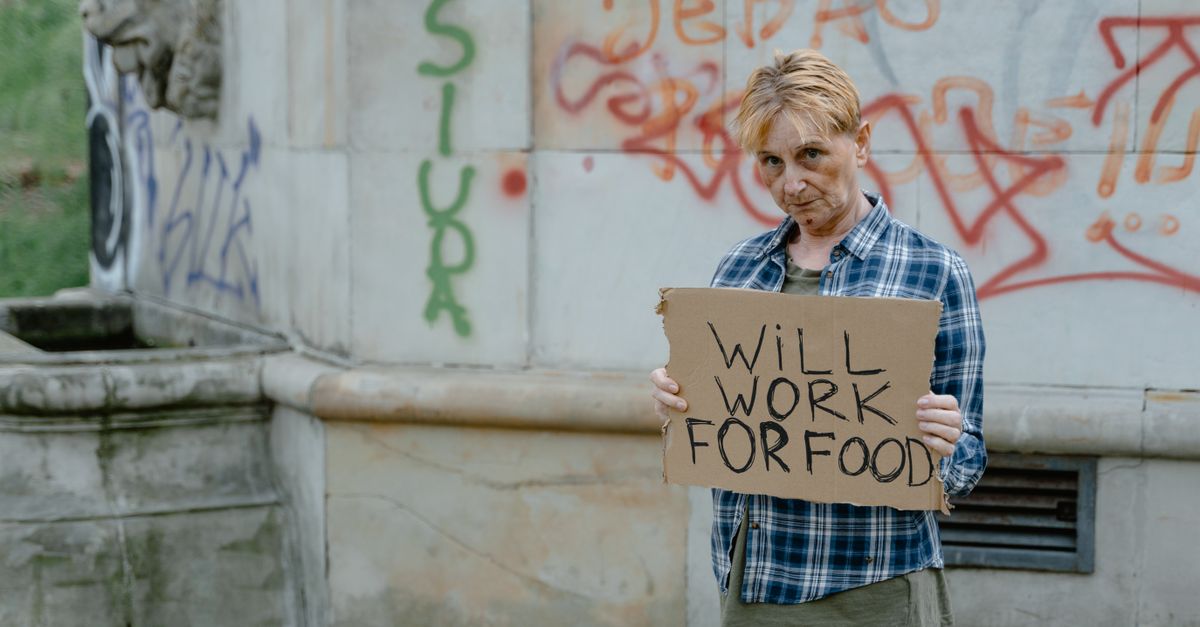 A homeless woman standing outdoors holding a sign that reads 'Will Work For Food'.