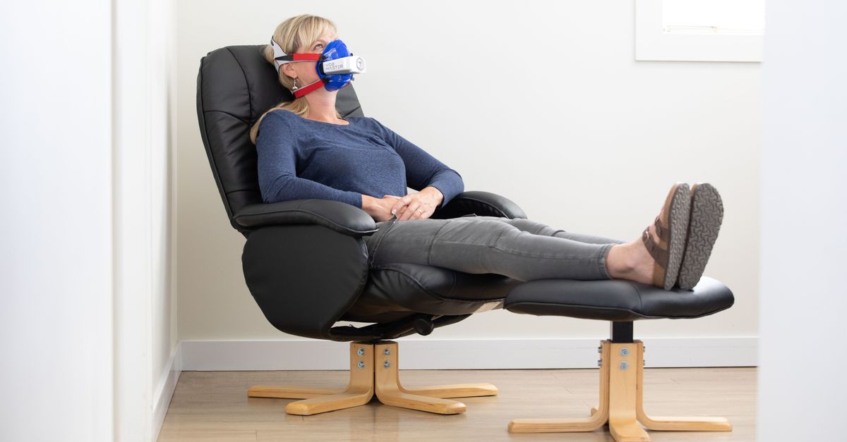 A woman comfortably reclines in a modern armchair, using a protective face mask indoors.
