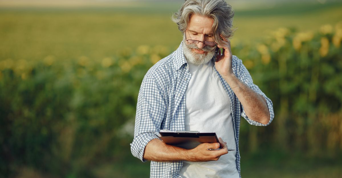 Elderly man in a field using a smartphone and tablet, wearing glasses and a casual shirt.