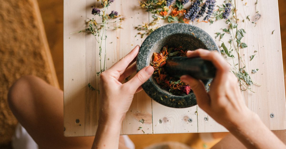 Hands using a mortar and pestle to grind colorful dried herbs on a wooden table.