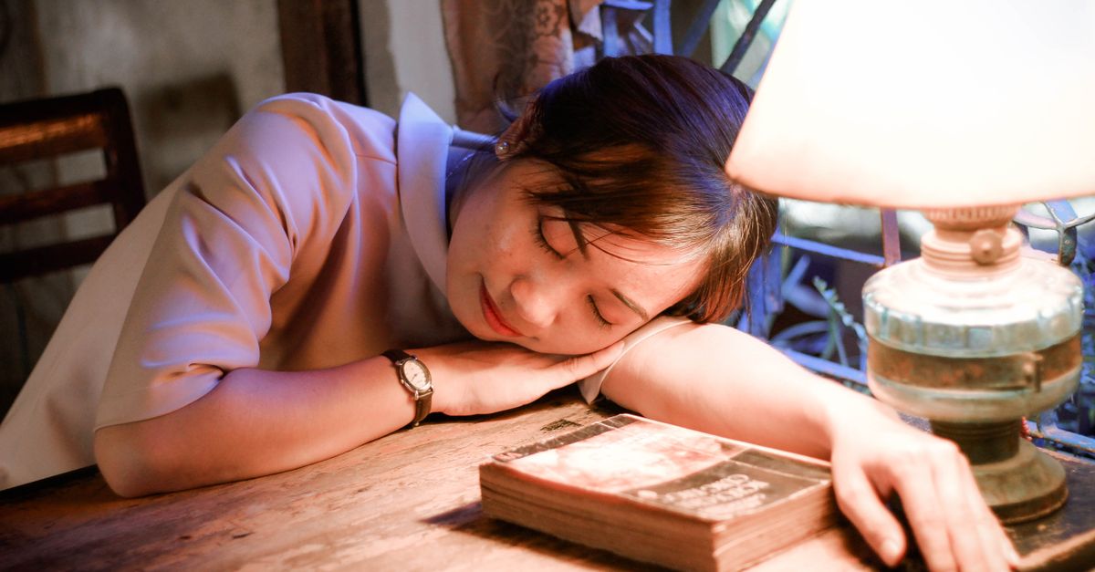 A young woman resting her head on a table under the warm glow of a lamp.