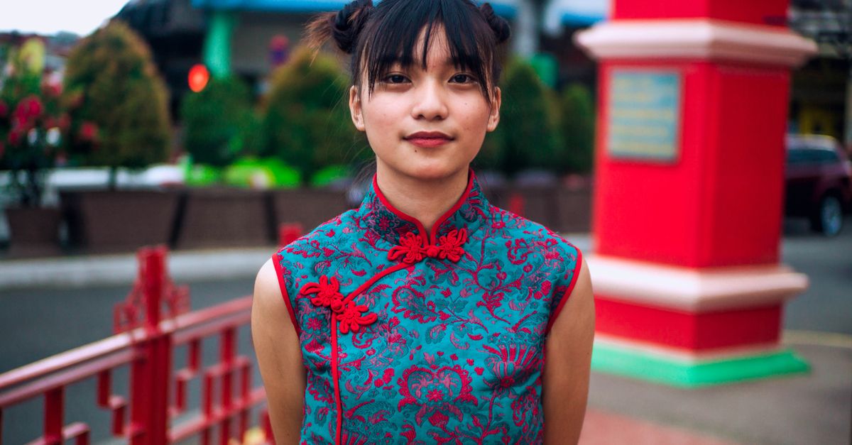 Positive young Asian female with trendy pigtail buns in traditional clothes chilling in city park and looking at camera