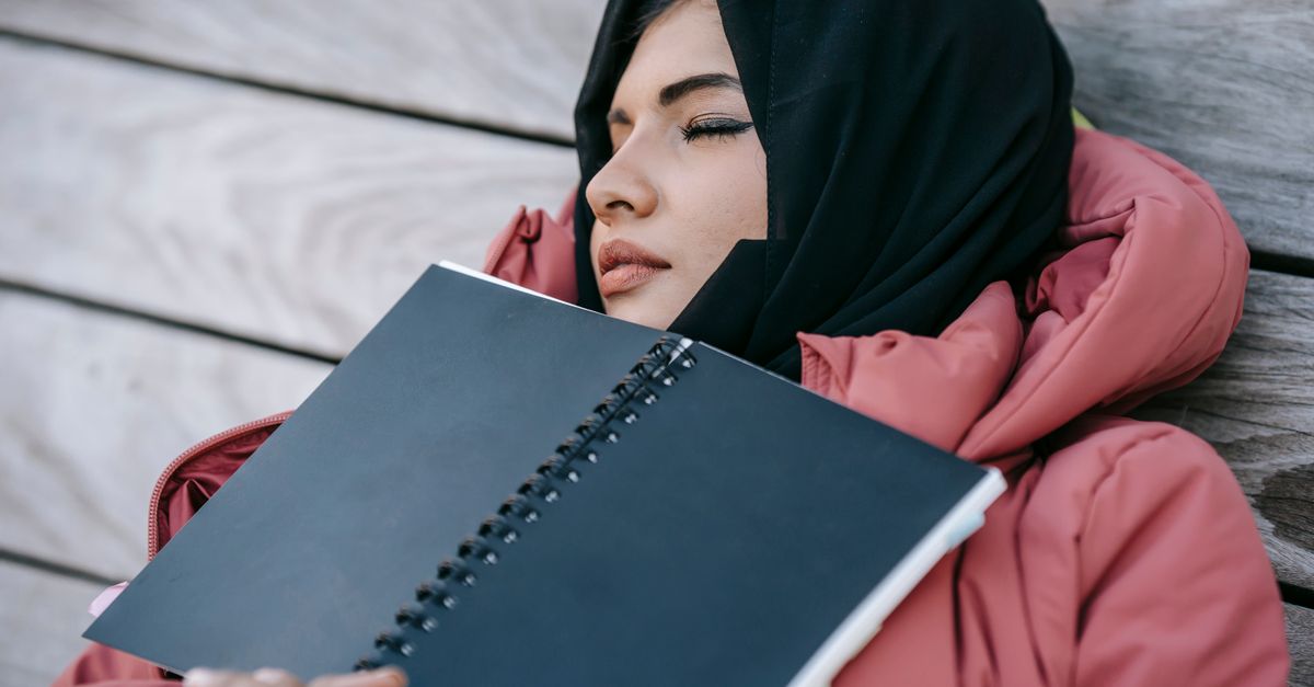 Young lady in hijab and warm outfit napping on wooden bench with notepad on chest in park