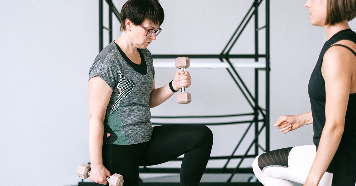 Elderly woman lifting dumbbells with guidance from a trainer indoors.