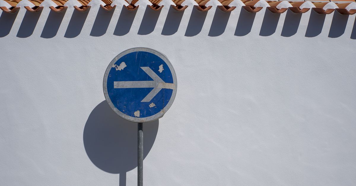 Blue arrow traffic sign casting shadow on a white wall with terracotta tiles.