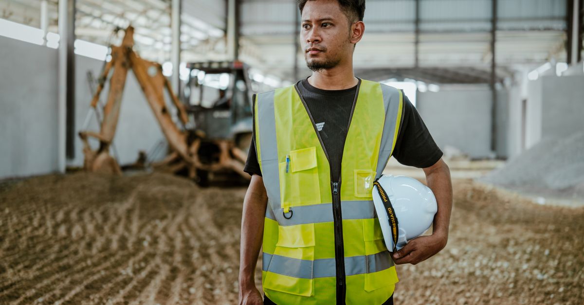 Portrait of a construction worker in safety gear holding a helmet inside an industrial setting.