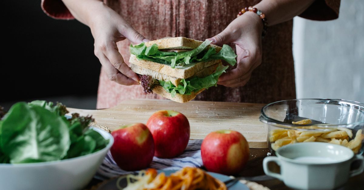 Unrecognizable female standing at table with apples and cutting board with sandwich with lettuce in hands in kitchen on breakfast time