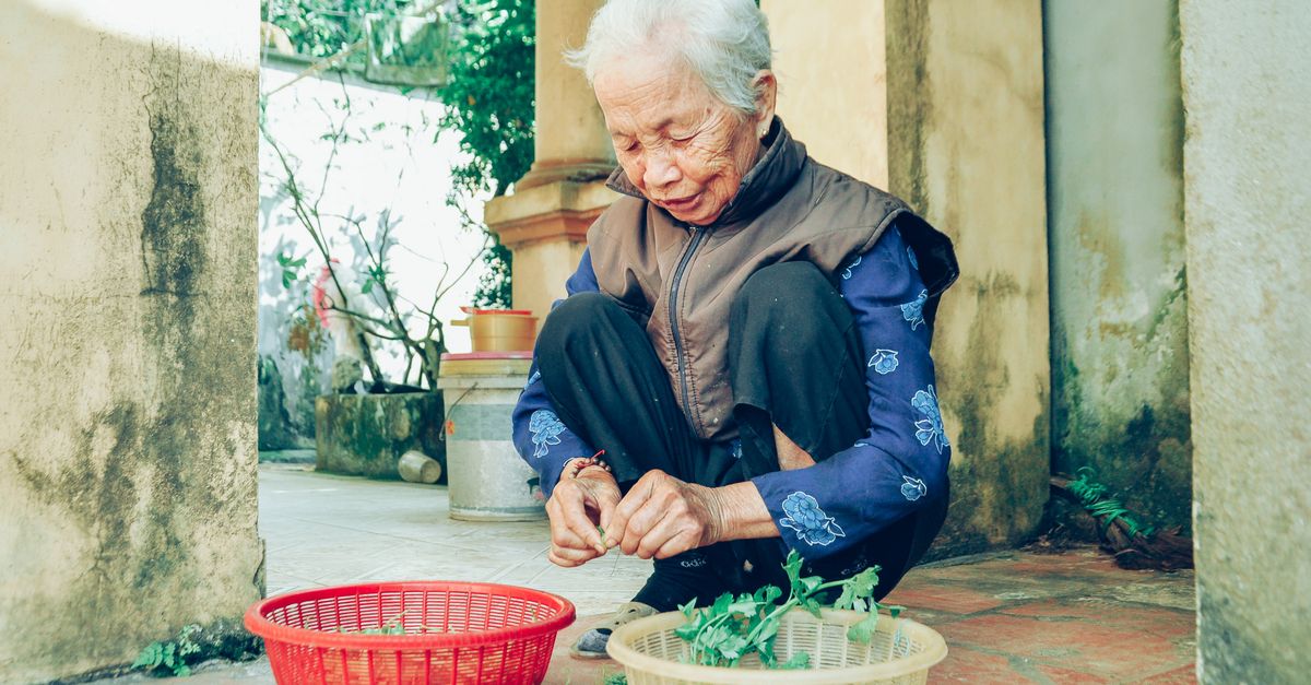 A senior woman sits outdoors, processing vegetables in baskets on a sunny day.