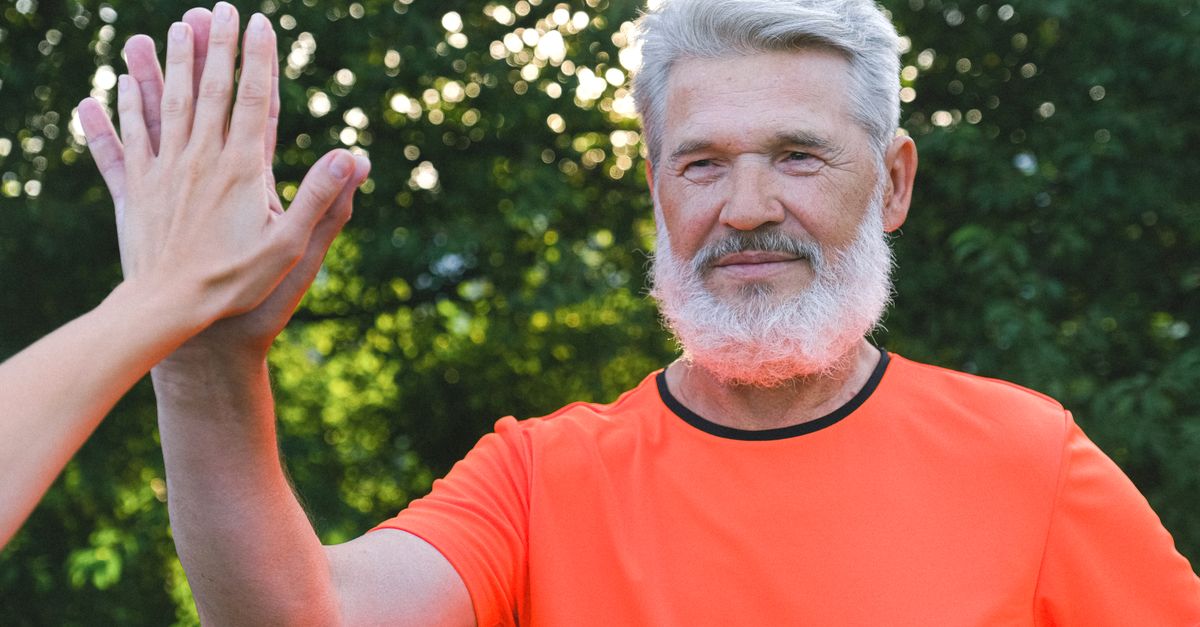 Calm aged man with beard in bright t shirt giving high five to anonymous person in park in sunny day