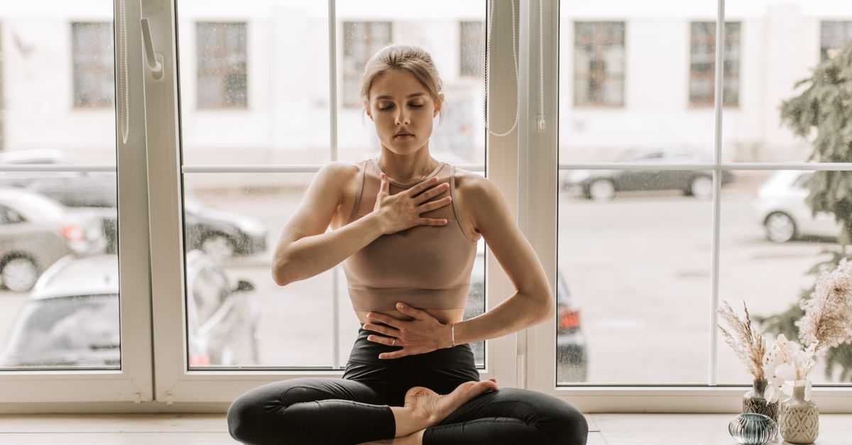 A woman practicing meditation in a serene yoga pose by a large window.