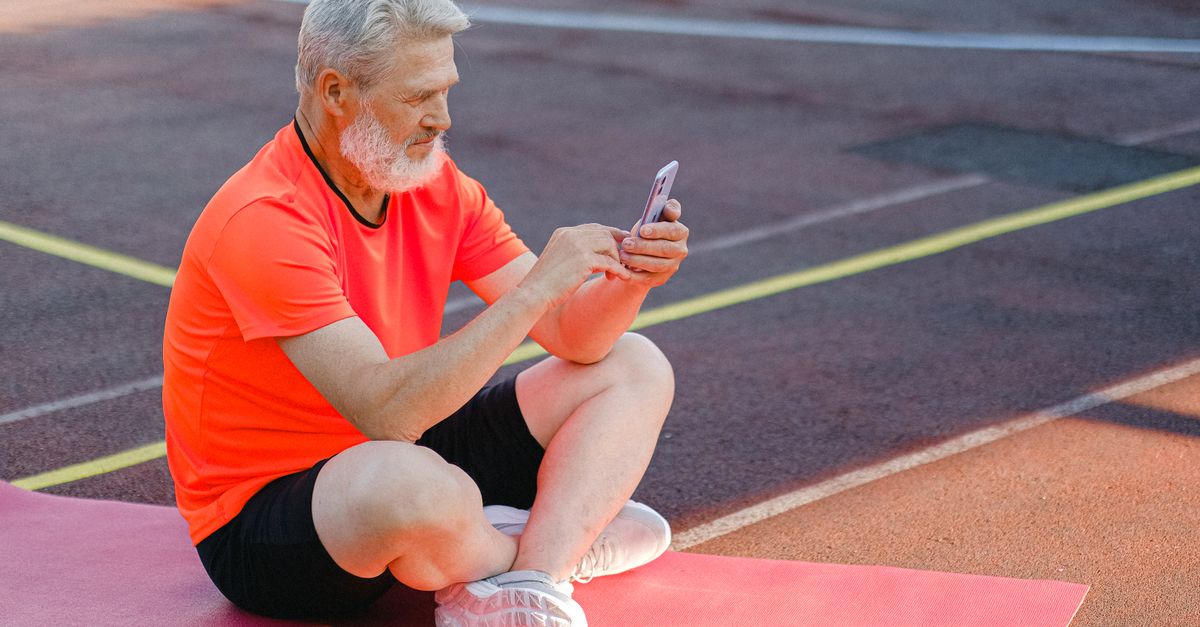 Full body of focused senior male athlete in sportswear sitting on stadium and browsing mobile phone during training